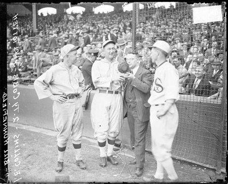 Baseball players, Eddie Collins, Ty Cobb, Bill Hunnefield, White Sox, and sportscaster, Hal Totten, of WMAQ radio with microphone in front of grandstands on the field at Comiskey Park